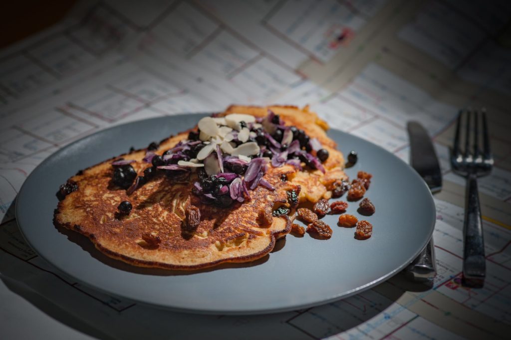 brown pancake with brown purple white nut on grey round plate beside silver knife and fork