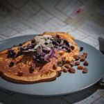 brown pancake with brown purple white nut on grey round plate beside silver knife and fork