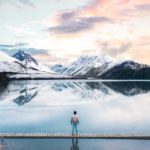 back view of a person standing on wooden planks across the snow capped mountains